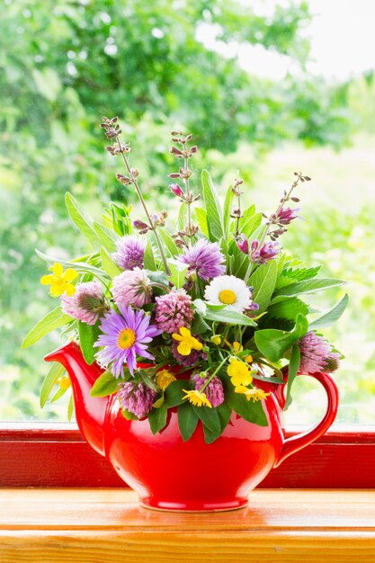 Tetera roja con ramo de hierbas curativas y flores en el alféizar de la ventana