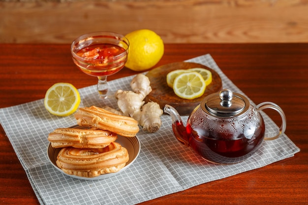 Una tetera de té caliente fuerte en una mesa de madera sobre una servilleta junto a limón, jengibre y mermelada y donas.