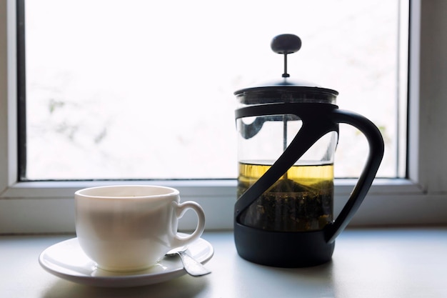 Tetera con té de hierbas y una taza blanca en el alféizar de la ventana. Bebida cálida y acogedora durante la estación fría. De cerca.