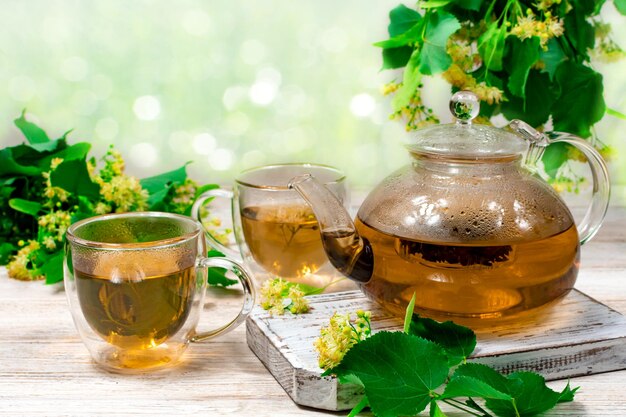 Una tetera de vidrio y dos tazas de té con un tilo sobre una mesa de madera sobre un fondo de hojas y flores de tilo Té curativo