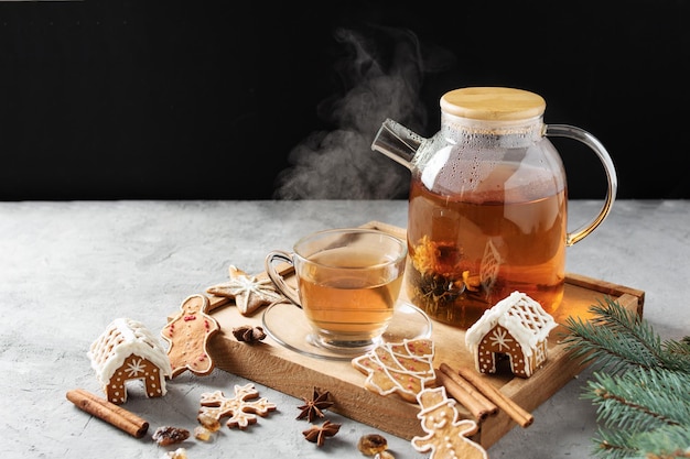 Tetera de vidrio con té floreciente y taza de té en una mesa gris con galletas de jengibre navideñas