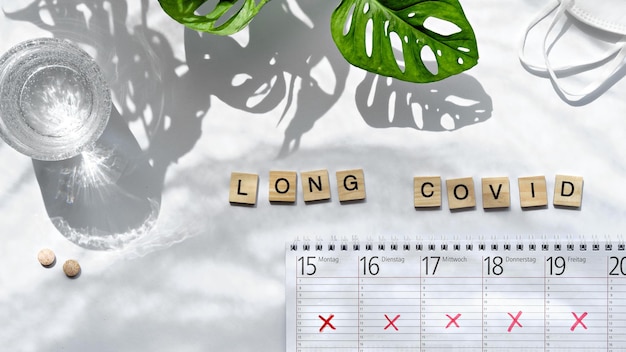 Text Long Covid, Holzbuchstaben. Wochenplaner, Kalender mit durchgestrichenen Tagesplänen. Absage, Planänderung wegen chronischer Covid-Erkrankung. Weißer Tisch mit Monstera-Blättern.