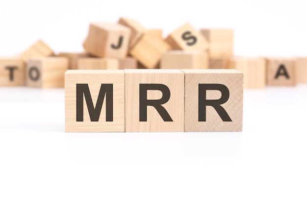 Text MRR Monthly Recurring Revenue é escrito em três cubos de madeira em pé sobre uma mesa branca ao fundo uma montanha de cubos de madeira com letras