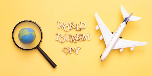Text zum Welttourismustag aus Holzbuchstaben auf gelbem Hintergrund mit Globus und einer Lupe und Flugzeugdraufsicht flach gelegt
