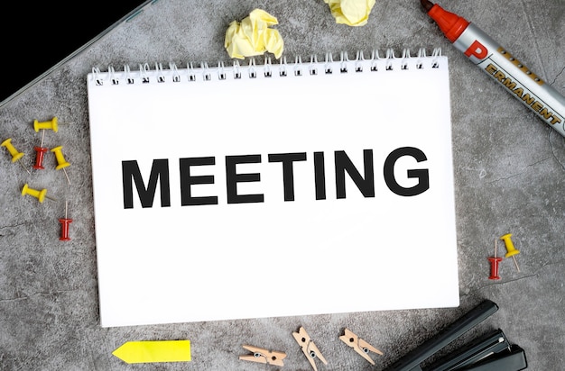 Texto de reunião em um caderno branco com alfinetes, marcador e grampeador em uma mesa de concreto