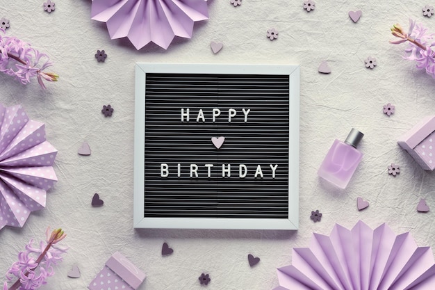 Texto feliz aniversário no quadro de cartas, papel timbrado. Postura plana criativa, vista superior em marfim, tecido branco. Decoração de festa, leques de papel, caixas de presente, corações. Flores de jacinto roxo claro natural, perfume.