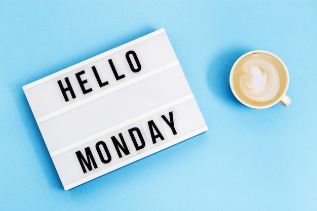 Texto "hola lunes" en caja de luz y taza de café para vacaciones. Comienzo del concepto de semana laboral. Vista superior sobre fondo azul.