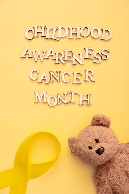 Foto texto del mes de concientización sobre el cáncer infantil con cinta y oso de peluche sobre fondo amarillo formato vertical