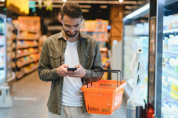 Texto rápido durante las compras Un joven apuesto sosteniendo un teléfono móvil y sonriendo mientras está de pie en una tienda de alimentos