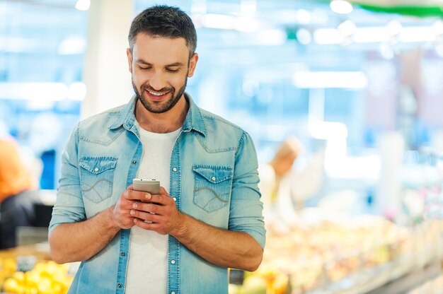 Foto texto rápido durante as compras. jovem bonito segurando um telefone celular e sorrindo enquanto está em uma loja de alimentos