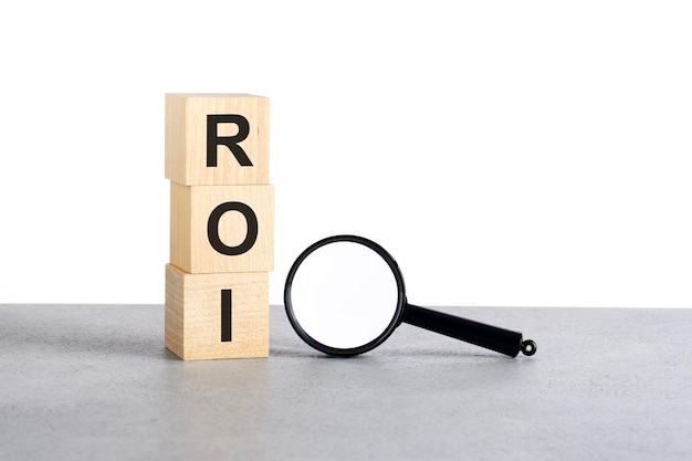 Texto de ROI Return On Investment en bloques de cubos de madera y lupa en mesa gris