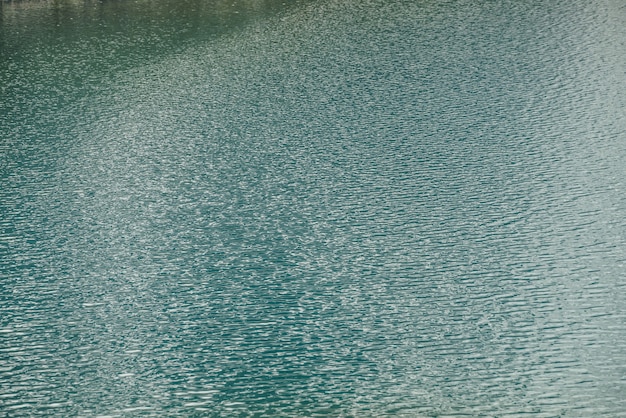 Textur des grün-blauen ruhigen Wassers des Sees. Meditative Wellen auf der Wasseroberfläche. Natur minimaler Hintergrund des grünblauen Sees mit goldenem Glanz. Natürliche Kulisse des Seefragments mit klarem azurblauem Wasser.