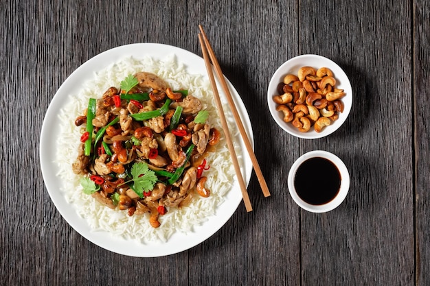 Thai-Cashew-Huhn-Pfanne, serviert mit Jasminreis auf einem weißen Teller auf einem Holztisch mit Stäbchen, horizontale Ansicht von oben, Nahaufnahme