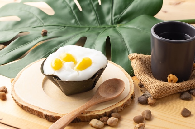 Thai Pudding Tapa de coco con semilla de Ginkgo biloba, Postres envueltos en hojas de plátano. Los tailandeses llaman "Khanom Tako". Tako con Ginkgo colocado en un platillo de madera y cuchara de madera, con té en vidrio negro.