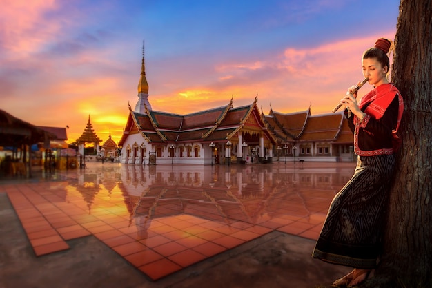 Thailändisches Mädchen in einer Nationaltracht, die zur thailändischen Flöte im buddhistischen Tempelbereich, Sakonnakhon, Thailand steht.