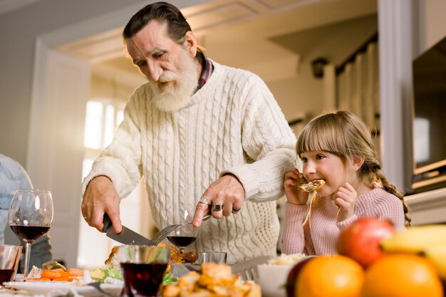 Thanksgiving-Feier Tradition Familiendinner-Konzept. Familie, die Feiertagsessen hat und Truthahn schneidet. Alter hübscher Großvater und seine Enkelin