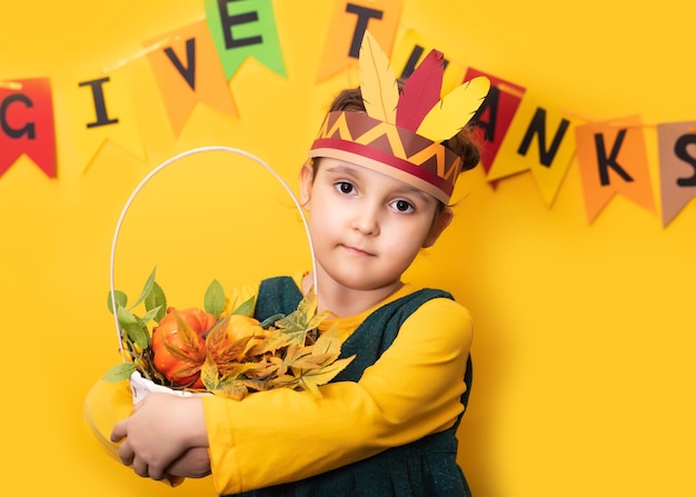 Thanksgiving-Tag-Konzept Süßes kleines Mädchen in Papierhut der amerikanischen Ureinwohner hält Korb mit Erntegeschenken auf gelbem Hintergrund