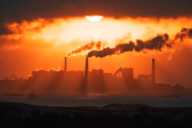 Thermoelektrische Schornsteine stoßen Dampf oder Rauch in einem feurigen Sonnenuntergang am Ufer des Ozeans aus
