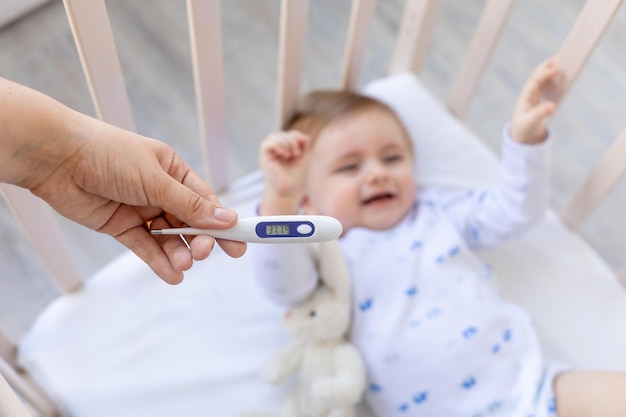 Thermometer im Fokus in Mamas Händen mit einer hohen Temperatur von 37 Grad auf dem Hintergrund eines kranken weinenden Jungen in einer Krippe zu Hause auf weißer Bettwäsche Medizin und Pädiatrie