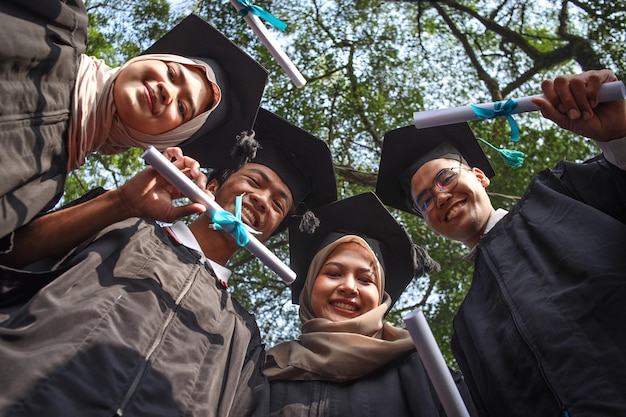 Tiefer Blickwinkel auf eine vielfältige Gruppe glücklicher junger Freunde, Studenten, die Abschlussroben tragen und halten