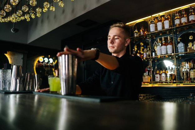 Tiefwinkelansicht eines jungen Barkeepers in schwarzer Kleidung, der einen erfrischenden alkoholischen Cocktail zubereitet, der hinter der Bartheke in einem modernen dunklen Nachtclub steht, auf dem Hintergrund von Regalen mit verschiedenen alkoholischen Getränken