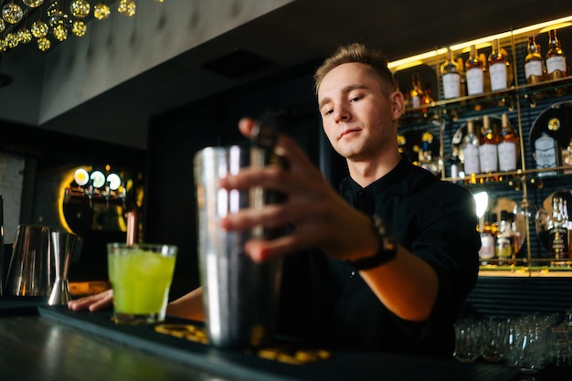 Tiefwinkelansicht eines selbstbewussten Barkeepers, der hinter der Bartheke in einem modernen dunklen Nachtclub bunte alkoholische Cocktails zubereitet