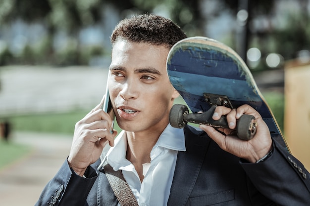 Tiempo de andar en patineta. Estudiante masculino agradable sosteniendo el monopatín y hablando por teléfono