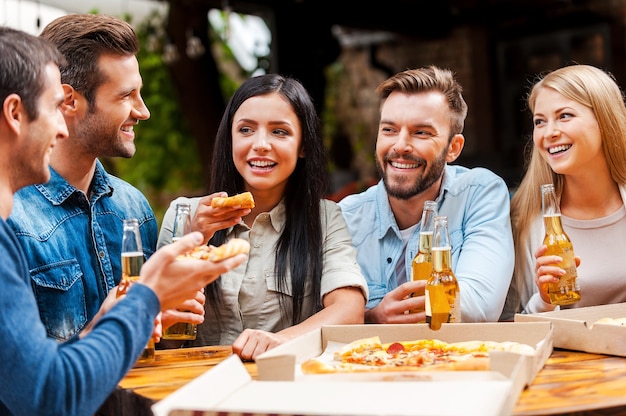 Tiempo de calidad con amigos. Grupo de jóvenes felices comiendo pizza y bebiendo cerveza mientras está de pie al aire libre