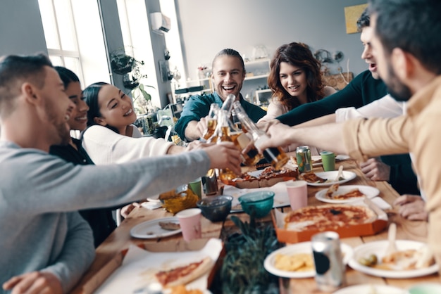 Tiempo de calidad entre amigos. Grupo de jóvenes en ropa casual brindando y sonriendo mientras tienen una cena en el interior