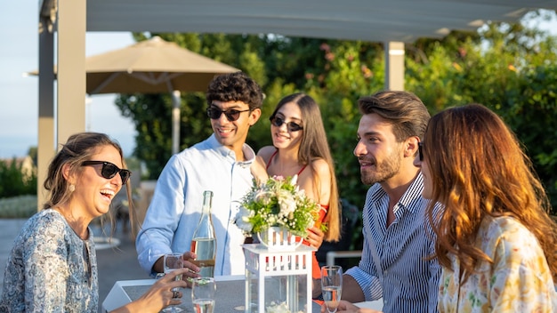 Tiempo de celebración jóvenes divirtiéndose fiesta al aire libre en la puesta de sol grupo de amigos sonriendo y riendo bebiendo vino blanco y brindando