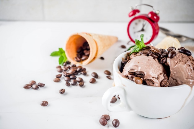 Tiempo para el concepto de café, con un despertador durante horas en el marco. Helado de café casero, servido con granos de café y hojas de menta, con conos de helado, cucharas. Fondo de mármol blanco, arriba