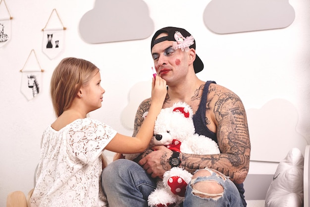 Tiempo divertido padre tatuado en una gorra y su hijo están jugando en casa linda chica está maquillando a él ...