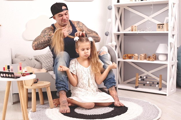 Tiempo divertido padre tatuado con una gorra y su hijo están jugando en casa papá está peinando a sus hijas ...