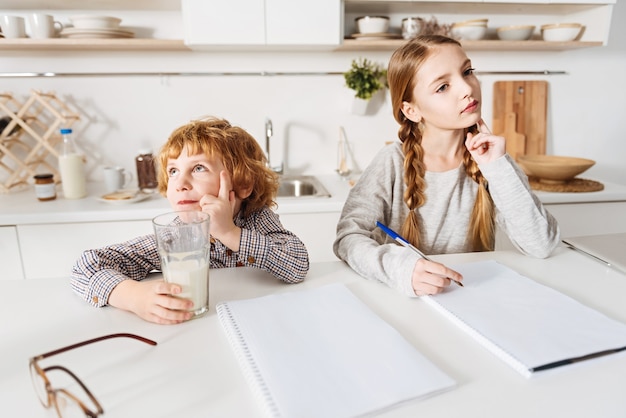 Tiempo de ensueño. Chica guapa inteligente responsable trabajando en su tarea en casa y su hermano pequeño tomando un vaso de leche mientras ambos se sientan en la cocina