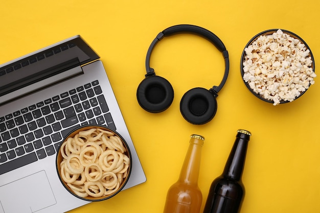 Tiempo de entretenimiento. Portátil con auriculares y bol de aperitivos, botellas de cerveza sobre fondo amarillo. Vista superior