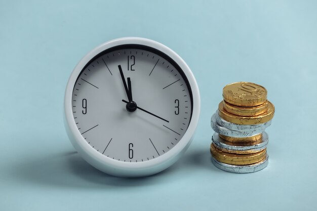 El tiempo es dinero. Reloj blanco con una pila de monedas sobre un azul