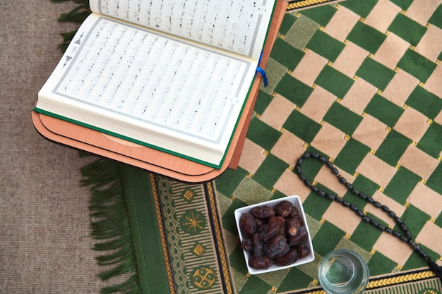 Tiempo de iftar Dátiles secos, vaso de agua del Sagrado Corán y tasbih sobre alfombra de oración o sejadah. Elementos que representan el mes de ayuno, el concepto de Ramadan Kareem