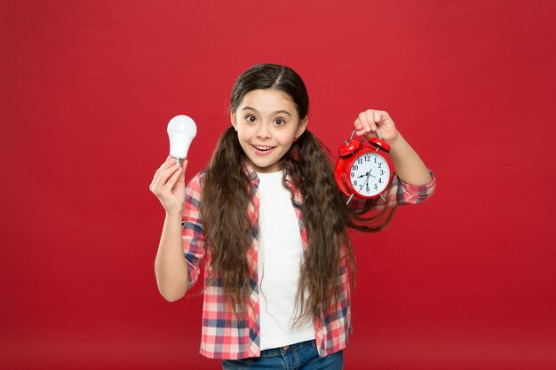 Tiempo de iluminación. Idea e inspiración. Niño inspirado mantenga fondo rojo del reloj. Inspiración y lluvia de ideas. Energía eléctrica. Bombilla de luz de retención de niña. Niño lindo en busca de inspiración.