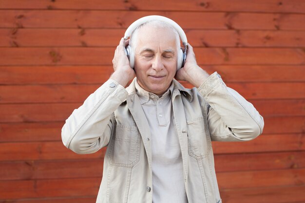 Tiempo de música relajante. Retrato de hombre senior en auriculares escuchando música manteniendo los ojos cerrados mientras está de pie contra el fondo de pared de madera