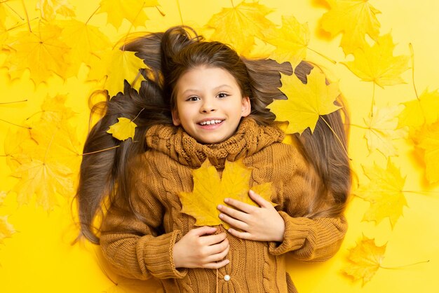 Tiempo de otoño Una niña en un suéter de punto yace sobre un fondo amarillo con hojas de arce caídas y agarra una hoja de arce en su pecho