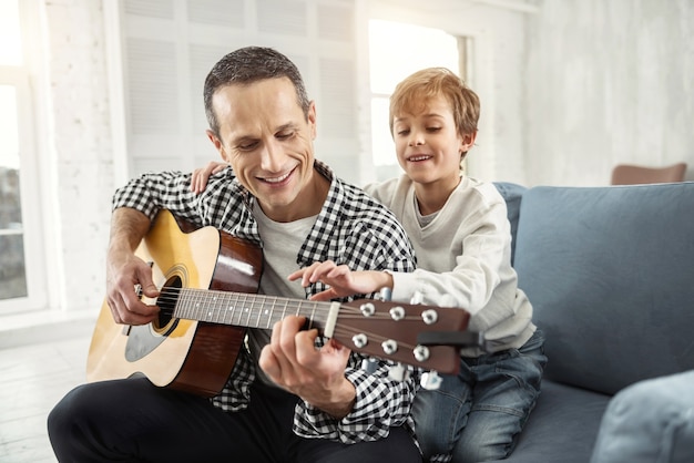 Foto tiempo precioso. apuesto hombre de pelo oscuro alegre sonriendo y tocando la guitarra y su hijo sentado detrás de él en el sofá