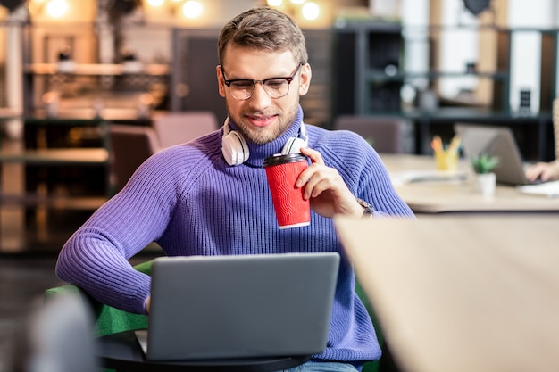 Tiempo para relajarse. Hombre barbudo complacido manteniendo una sonrisa en su rostro mientras usa su computadora portátil