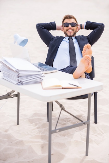 Foto tiempo para relajarse. hombre joven alegre en ropa formal y gafas de sol tomados de la mano detrás de la cabeza y sosteniendo sus pies sobre la mesa de pie sobre la arena