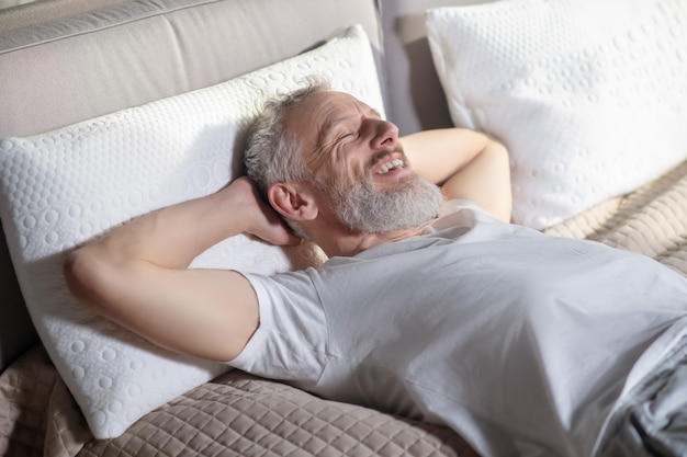 Foto tiempo de relax. hombre canoso con barba acostado en la cama con las manos levantadas detrás de la cabeza sonriendo con los ojos cerrados
