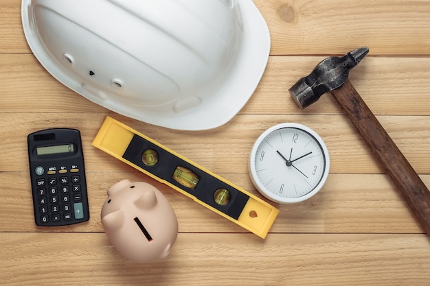 Tiempo de reparación. Casco de seguridad de construcción blanco, martillo, reloj, alcancía y calculadora, nivel sobre tablas de madera. Vista superior. Costos de reparación. Idea de construcción