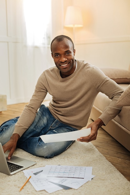 Tiempo de trabajar. Sonriente hombre afroamericano barbudo de ojos oscuros sonriendo y trabajando en la computadora portátil y sosteniendo una hoja de papel mientras está sentado en el piso y otros papeles que yacen cerca de él