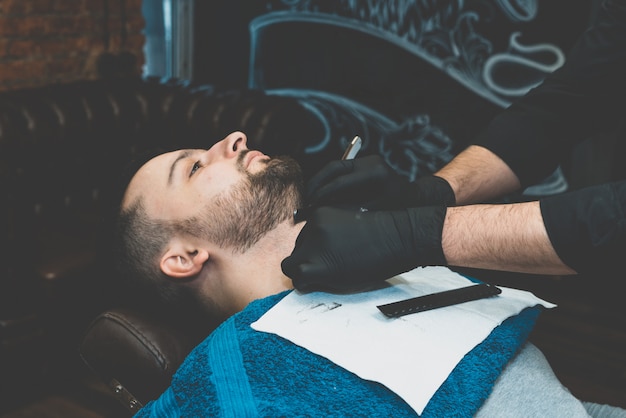 Tienda de belleza para hombres. Afeitarse la barba en una barbería. Barber se corta la barba con una navaja y una maquinilla. Cerrar cortes de pelo brutales. Equipamiento de peluquería. Enfoque selectivo.