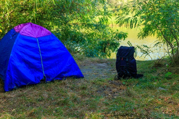 Tienda de campaña azul y mochila en el bosque cerca de un río