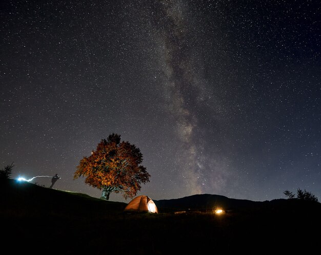 Tienda de excursionistas turísticos iluminada desde el interior, silueta de hombre y fogata ardiente bajo el cielo estrellado azul oscuro.