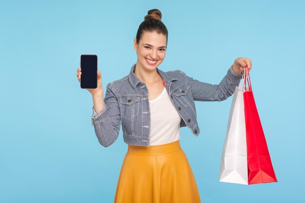 Tienda en línea. Retrato de mujer atractiva vestida a la moda con moño de pelo sonriendo, sosteniendo paquetes y teléfono celular, publicidad de compras móviles. Foto de estudio interior aislado sobre fondo azul.